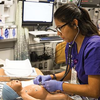 一名护理bc体育的学生穿着紫色的手术服，在一个婴儿模拟假人上练习听诊器