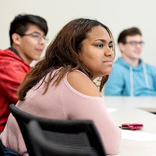 三名不同种族的TCU学生在教室里一起听讲. 前景是一位身穿粉红色毛衣的年轻女子.